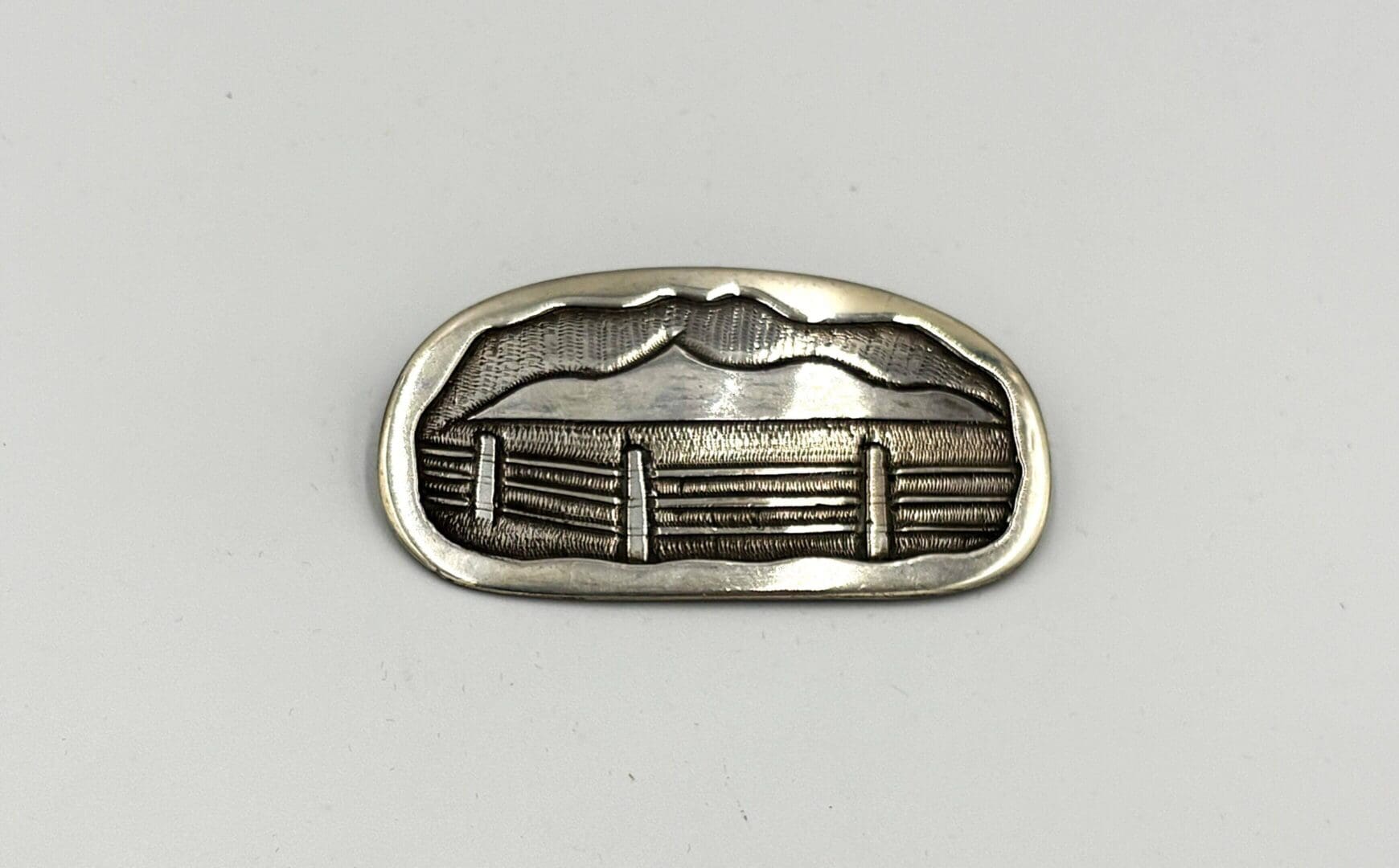 Oval metal piece with engraved mountain scene.